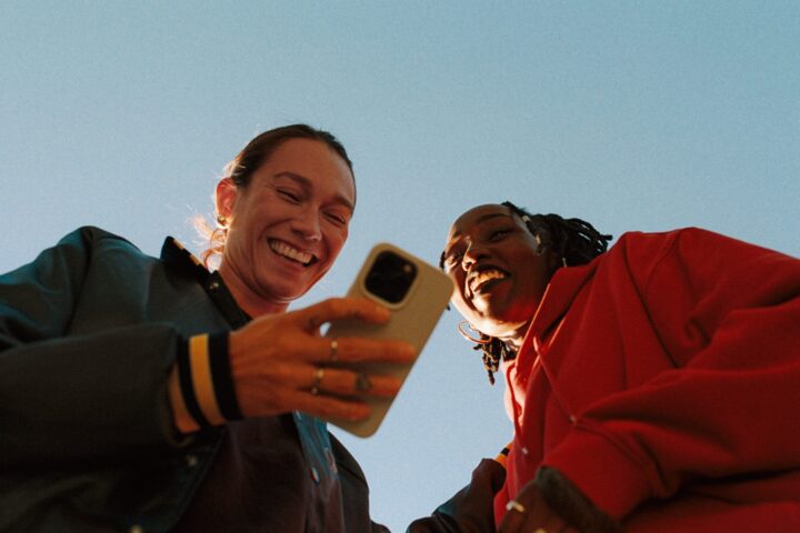 Två unga leende kvinnor står utomhus och tittar i en smartphone.