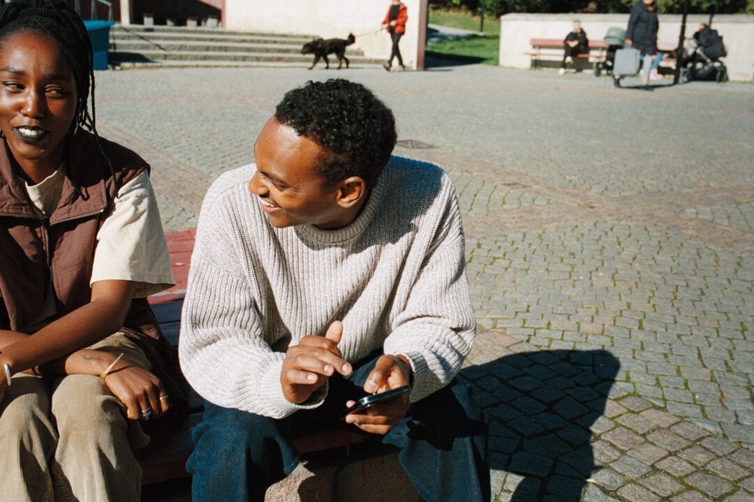 Ung kvinna och man sitter utomhus på en bänk i solen.
