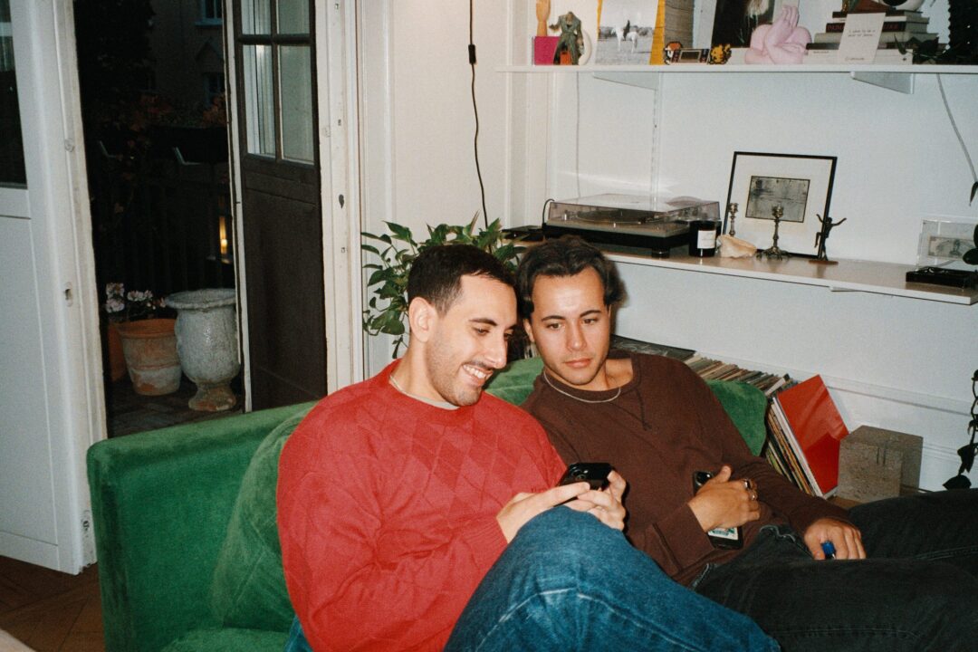 Två män sitter i en soffa i en lägenhet. Den ena håller i en mobiltelefon.