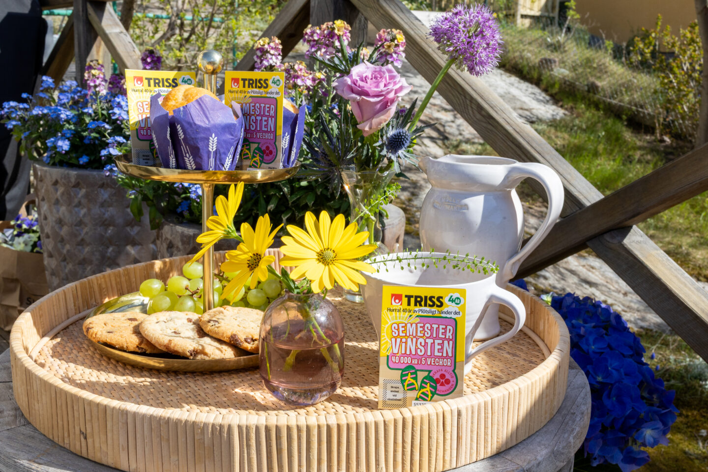 Tre Triss Semestervinsten-lotter på ett dukat sommarbord med blommor, kakor, vindruvor och kaffekopp i trädgårdsmiljö.